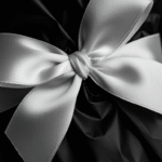 An extreme close-up of a beautifully tied white satin ribbon forming a bow atop a bouquet wrapped in black paper. The satin texture is smooth and shiny, with the bow's loops and tails clearly defined. The black wrapping paper shows a sharp fold, creating a dramatic contrast with the ribbon. The shallow depth of field blurs the background, emphasizing the detailed texture of the satin and the crispness of the folding paper.