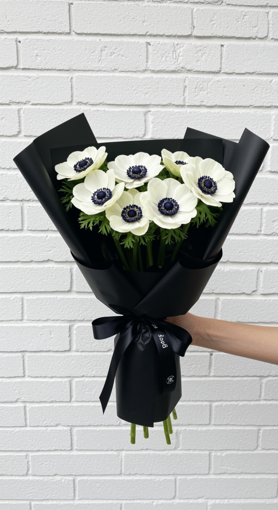 A close-up of a bouquet of white anemones with prominent black centers, wrapped in black paper and tied with a thin black ribbon, held against a white brick wall background, highlighting the striking contrast between the flowers and the backdrop.