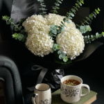 A beautifully arranged bouquet of cream hydrangeas and sprigs of eucalyptus, wrapped in elegant black paper, rests on a plush velvet armchair. Next to it, a steaming mug and an open book add a cozy, inviting touch. The scene is styled to evoke warmth and sophistication, perfect for a lifestyle or floral decor setting.