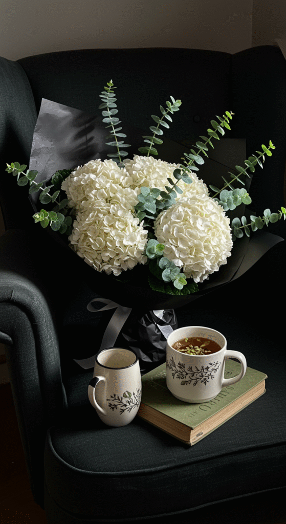 A beautifully arranged bouquet of cream hydrangeas and sprigs of eucalyptus, wrapped in elegant black paper, rests on a plush velvet armchair. Next to it, a steaming mug and an open book add a cozy, inviting touch. The scene is styled to evoke warmth and sophistication, perfect for a lifestyle or floral decor setting.
