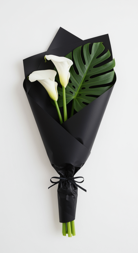 A minimalist bouquet featuring three white calla lilies and a single large monstera leaf, wrapped in a sleek black paper, set against a plain, uncluttered background