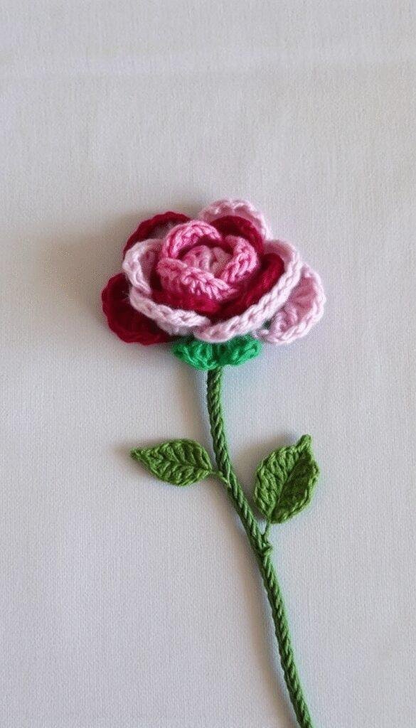 A close-up of a vibrant, handcrafted crochet rose flower in shades of pink and red, balanced on a soft white background with detailed yarn textures visible, showcasing intricate stitches and realistic petal shapes.