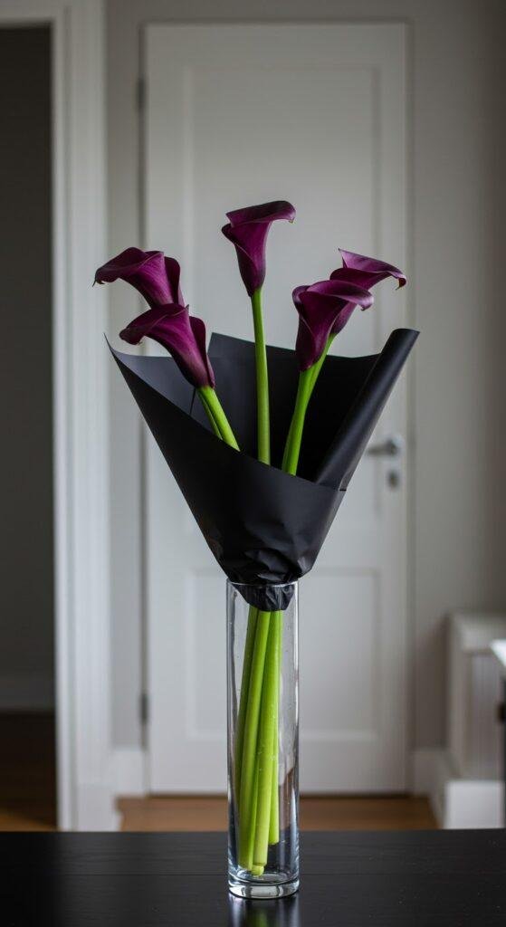 A sleek, architectural bouquet of five long-stemmed deep purple calla lilies, wrapped in black paper highlighting their elegant form, placed in a tall, slender glass vase on a minimalist tabletop in a contemporary apartment.