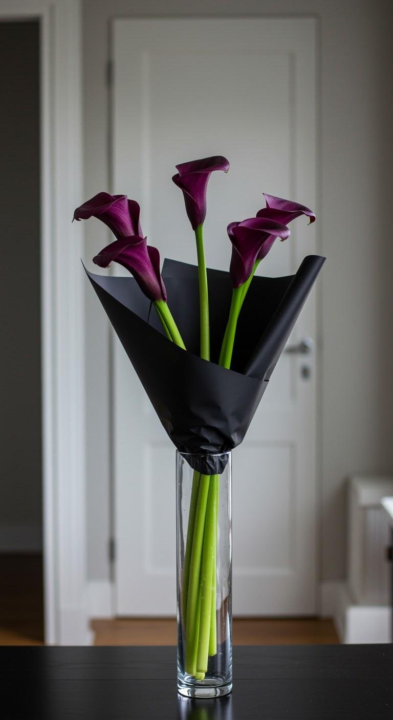 A sleek, architectural bouquet of five long-stemmed deep purple calla lilies, wrapped in black paper highlighting their elegant form, placed in a tall, slender glass vase on a minimalist tabletop in a contemporary apartment.