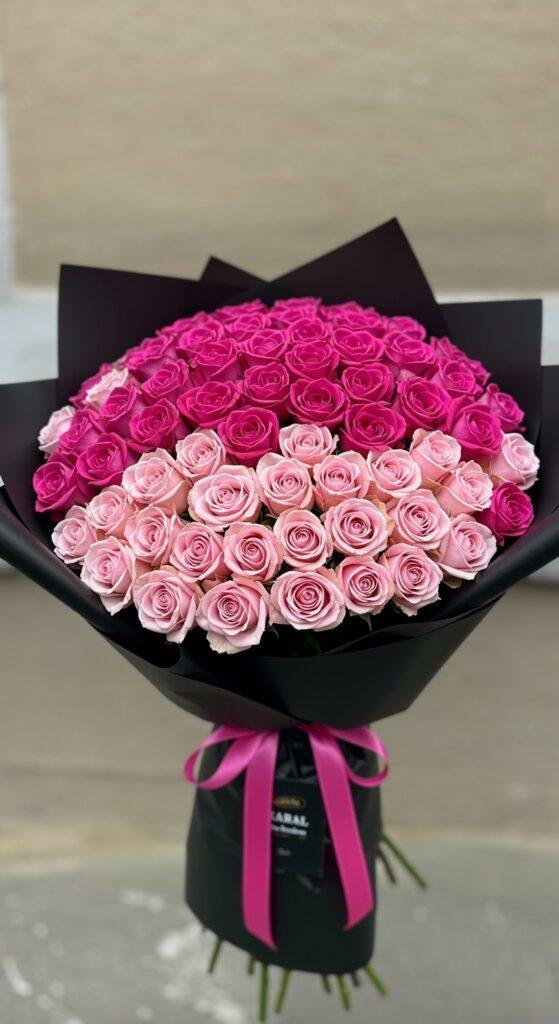 A stunning bouquet of roses arranged in an ombré gradient, transitioning from deep magenta at the top to pale pink at the bottom, wrapped in black paper to enhance the vibrant colors. The bouquet is photographed against a neutral, softly blurred background, highlighting the beauty of the color transition and the elegant floral arrangement.