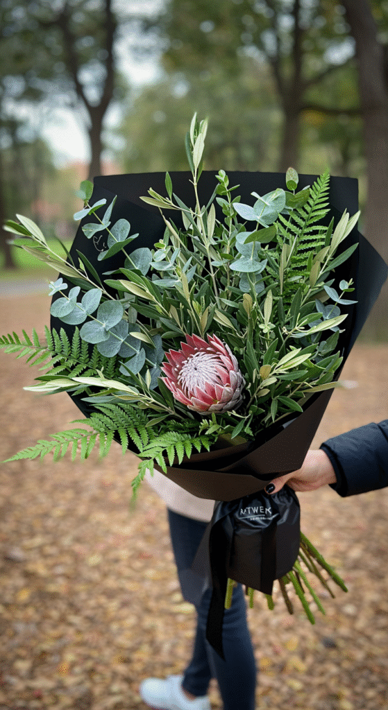 A lush, textured bouquet composed entirely of various eucalyptus leaves, ferns, and olive branches wrapped in black paper. The bouquet is being held by a person walking through a park, with greenery and trees in the background, creating a vibrant, nature-inspired, chic vibe.