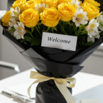 A vibrant bouquet of yellow roses and white alstroemeria arranged professionally and wrapped in black paper, placed on a sleek office desk with a welcoming 'Welcome' card beside it, illuminated by bright natural light.