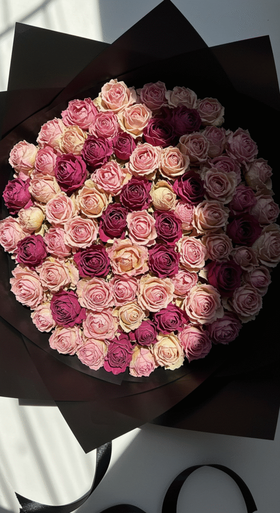 A flat lay image of dried rose petals arranged in an artistic pattern on a neutral surface, with the original black wrapping paper and ribbon placed nearby, creating a sophisticated floral display